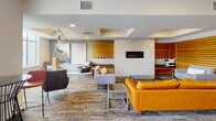 Located at 519 Porter St in Richmond, Virginia, this single-floor communal space offers a thoughtfully designed environment with distinct zones for relaxation, socializing, and casual gathering. The layout flows openly between areas, anchored by a central lounge featuring a linear gas fireplace framed by warm wood slatted wall panels. Seating throughout includes a mix of leather sofas, modern accent chairs, and upholstered benches arranged across patterned carpet flooring, creating an inviting and contemporary atmosphere.

A dedicated game area accommodates leisure activities, while pendant light fixtures with geometric metal frames add a sophisticated design touch overhead. Large-scale framed artwork adorns the walls, contributing to the polished aesthetic. A café-style seating area near floor-to-ceiling glass doors and expansive windows provides panoramic views of the Richmond skyline, with direct access to an outdoor terrace equipped with bistro-style seating.

The kitchenette area features sleek gray cabinetry with under-cabinet lighting, white countertops, a stainless steel sink, and light tile flooring. An accessible restroom is conveniently located nearby. Recessed lighting and drop ceilings throughout ensure consistent illumination across the entire space.