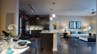 This single-floor space showcases a thoughtfully designed open-concept layout that integrates the kitchen, dining, and living areas into one cohesive environment. The kitchen is a standout feature, outfitted with rich dark cabinetry that extends to the ceiling, providing ample storage. Granite countertops add a touch of elegance, while a central island with bar seating serves as both a functional workspace and a social gathering point. Pendant lighting hangs above the island, complementing the track lighting overhead to create a well-lit, layered ambiance. Stainless steel appliances, including a refrigerator and stove, complete the modern kitchen aesthetic. A subtle tile backsplash adds texture between the countertops and upper cabinets. Dark hardwood flooring runs throughout, providing a cohesive and sophisticated foundation. The open sightlines from the kitchen extend naturally into the dining and living areas, where teal accents in furnishings introduce a pop of color against the neutral palette. A doorway visible from the kitchen suggests additional rooms beyond, hinting at a well-connected and functional floor plan throughout the space.