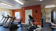 This dedicated fitness space is thoughtfully designed to support a full range of workout activities. The room features bold terracotta-orange accent walls paired with neutral grey ceilings, creating an energizing and motivating atmosphere ideal for exercise. Durable dark rubber flooring covers the entire space, providing excellent traction and impact absorption during workouts. The room is well-lit through a combination of overhead fluorescent lighting and natural light from a window, keeping the environment bright and functional. A variety of cardio equipment is available, including a recumbent bike and an elliptical cross-trainer, catering to different fitness preferences and intensity levels. A weight rack stocked with a full set of dumbbells is positioned along one wall, offering strength training options. The layout is practical and spacious, allowing users to move freely between equipment. A standard door provides easy access in and out of the space. Overall, this fitness room is a fully functional, purpose-built environment that delivers everything needed for both cardiovascular and strength-based training routines.