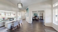 This single-story community clubhouse is thoughtfully designed to serve as a central hub for residents, offering a variety of amenity spaces within a bright, welcoming layout. The entrance features a decorative glass-panel front door with transom windows that flood the foyer with natural light, complemented by wood-look plank flooring that flows consistently throughout the building.

The leasing office area includes a clean white reception counter with vaulted ceilings and a pendant light fixture, with windows overlooking the indoor pool. French doors with glass panes connect the various rooms, creating an open and airy feel while maintaining defined spaces.

The lounge is a standout area, anchored by a full-height brick fireplace with a mounted flat-screen television and a built-in window bench. Adjacent to the lounge is a well-equipped kitchen bar featuring an elongated white cabinetry run with warm countertops, mirrored backsplash, pendant jar lighting, and a coffee station, ideal for resident gatherings.

A casual seating nook with a round table and blue chairs provides a relaxed setting, accompanied by a vending machine for convenience. The fitness center offers both cardio and strength-training equipment, set against bold blue accent walls with mirrored panels and carpeted flooring.