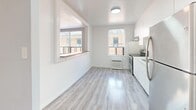 Located at 2305 Lake Shore Blvd W in Toronto's lakeshore neighbourhood, this one-bedroom, one-bathroom unit offers a clean, modern aesthetic throughout. Light grey wood-look flooring runs continuously across the entire space, creating a cohesive and airy feel. The open layout connects the living room to the combined dining and kitchen area, which is separated by a partial wall featuring a convenient pass-through window. The kitchen is well-appointed with white cabinetry, marble-effect countertops, a classic white subway tile backsplash, and stainless steel appliances including a refrigerator and range. Natural light flows in through multiple windows, complemented by classic radiator heating units beneath them. The spacious bedroom includes a closet and a window overlooking the surrounding neighbourhood, with ample room for a full furniture arrangement. The bathroom is finished with marble-effect wall and floor tiles, a soaking tub with a showerhead and curtain rod, and a white vanity. Two closets provide practical storage throughout the unit. The entrance leads directly into the main living area, offering a straightforward and functional flow across the single-floor layout.