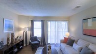 This single-story space presents a welcoming living room that serves as the heart of the home. The room is filled with natural light, courtesy of a large sliding glass door fitted with vertical blinds and an adjacent window dressed with elegant grey curtain panels. Together, these elements offer both abundant daylight and a connection to the outdoors.

The living area is thoughtfully arranged, featuring a neutral-toned sofa accented with warm coral and grey throw pillows, complemented by a coordinating accent chair in a muted blue-grey. A patterned area rug anchors the seating arrangement atop hardwood-style flooring, adding texture and visual interest to the space.

The walls are finished in a soft, warm neutral tone, while the textured ceiling adds subtle architectural character. Floor lamps on either side of the room provide layered ambient lighting, enhancing the overall warmth of the space.

The layout feels open and functional, with the sliding glass door providing easy access to the outdoors. The overall ambiance is calm and contemporary, with a cohesive color palette that gives the space a clean, move-in-ready feel.