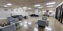 Located at 526 Superior Avenue in Cleveland, Ohio, this single-floor commercial space offers a thoughtfully designed environment suited for both relaxation and activity. The expansive main lounge area is anchored by polished terrazzo flooring that reflects the abundant overhead lighting, creating a bright and welcoming atmosphere. Comfortable seating arrangements — including curved sofas, upholstered armchairs, and glass-topped coffee tables — are artfully arranged atop decorative area rugs, encouraging conversation and collaboration. Framed artwork adorns the neutral-toned walls, adding a touch of character to the professional setting. Structural columns punctuate the open floor plan while defining distinct zones within the space. A raised area visible from the lounge provides additional seating and dining furnishings, enhancing the versatility of the layout. Large interior windows along one wall offer a clear view into a fully equipped fitness center, where a range of exercise machines is visible, seamlessly integrating wellness into the facility. The drop-panel ceiling with recessed and flush-mounted lighting ensures consistent, even illumination throughout. Overall, the space strikes a balance between professional functionality and comfortable, modern design.