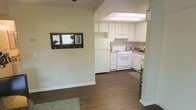 This single-floor space features a straightforward, functional layout with an open sightline connecting the main living area to the kitchen. The flooring throughout is dark hardwood, providing a warm contrast against the light-colored walls and white trim. A small, framed pass-through window with hooks beneath it is mounted on the living area wall, serving as a practical entryway organizer or decorative accent.

The kitchen is compact yet well-organized, featuring white upper and lower cabinetry that maximizes storage. A white gas range and a full-size white refrigerator are neatly positioned within the layout, complemented by light-toned countertops. Overhead, a flush panel light fixture provides bright, even illumination across the workspace. The kitchen transitions smoothly into the adjacent area, maintaining a cohesive flow throughout the space.

The overall ambiance is clean and neutral, with a simple, utilitarian aesthetic. Light-toned walls and white fixtures keep the interior feeling bright and open despite its modest footprint. A small area rug anchors the living area, adding a touch of softness to the hard flooring surfaces. The space offers a practical and easy-to-maintain environment suitable for everyday living.