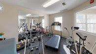 This single-floor space functions as a combined fitness and physical rehabilitation facility, thoughtfully outfitted to accommodate a range of exercise and therapeutic activities. The room features neutral-toned walls, carpeted flooring in select areas, and adequate overhead lighting supplemented by natural light streaming through multiple windows fitted with adjustable blinds.

The fitness area is well-equipped with an array of cardio machines, including a treadmill, stationary bikes, an air resistance bike, and an elliptical trainer. A road bicycle is also present, adding versatility to cardiovascular training options. Full-length mirrors line one wall, enhancing the sense of space and allowing users to monitor their form during workouts. Free weights, a barbell, resistance bands, and a stability ball are organized on a storage rack, providing a comprehensive selection of strength and flexibility training tools.

A dedicated rehabilitation zone features a professional-grade treatment table with a face cradle and an adjacent scale, suggesting clinical or therapeutic use. A frosted-glass interior door separates sections of the space, offering a degree of privacy. A fire alarm and security camera indicate attention to safety. The overall layout is functional and purposeful, supporting both independent fitness routines and structured rehabilitation sessions.