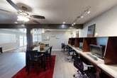 Located at 1501 Harvey Road in College Station, Texas, this single-floor community space offers a thoughtfully designed open layout that seamlessly blends a lounge area, a dining area, and a dedicated computer workstation zone. The interior is finished with durable wood-look flooring throughout, complemented by neutral gray walls and crisp white baseboards that lend a clean, modern aesthetic.

The lounge area welcomes visitors with comfortable seating, including a beige sofa and upholstered chairs arranged around a glass coffee table atop a colorful area rug. Bright windows with white blinds allow ample natural light to fill the space, while ceiling fans ensure year-round comfort. Framed artwork adorns the walls, adding a welcoming touch.

Transitioning deeper into the space, a dining area features a dark wood table with matching chairs set atop a bold red area rug, ideal for group meetings or casual gatherings. The workstation section is equipped with a long built-in desk, dark wood privacy dividers, multiple computer monitors, and ergonomic office chairs, all illuminated by track lighting overhead. A small seating nook with red accent chairs and wall-mounted shelving provides additional functionality. The space is well-lit, practical, and designed to serve as a productive shared environment.