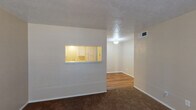 This single-floor residence offers a practical and functional layout designed for comfortable everyday living. The living area is finished with plush dark carpeting and neutral-toned walls, creating a warm and inviting atmosphere. A standout architectural detail is the pass-through window connecting the living space to the kitchen, allowing natural communication between the two areas while also brightening the space with the kitchen's warm lighting. The living room also opens into a dining or flex area featuring wood-look flooring, a ceiling-mounted light fixture, and a ventilation system, giving the space a clean and connected flow.

The kitchen is efficiently arranged with white cabinetry, an electric coil range, and a full-size refrigerator, all set on wood-look flooring. A small window above the range provides ventilation and a touch of natural light. The bedroom is a comfortable, well-sized retreat with soft carpeting, a ceiling fan with light for year-round comfort, and a window fitted with blinds for privacy and light control.

The bathroom is neatly appointed with a white vanity, framed mirror, lighted fixture above, additional wall-mounted cabinetry for storage, and wood-look flooring that ties the space together cohesively. Throughout the residence, a consistent use of neutral tones and practical finishes contributes to a clean, move-in-ready aesthetic.