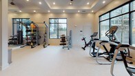 This fitness center is designed to support a comprehensive workout experience within a single, open-plan space. The room features a tray ceiling with recessed lighting that bathes the light-toned flooring in a warm, even glow, contributing to an inviting and energetic atmosphere. Large black-framed windows along multiple walls allow abundant natural light to fill the space while providing views of the surrounding urban environment.

The cardio zone is equipped with treadmills and an elliptical trainer, positioned near the windows to make use of natural light during workouts. Strength and functional training needs are addressed through a cable machine, a dedicated TRX suspension system, and a well-stocked rack holding medicine balls, resistance bands, and exercise mats. A flat bench and stationary bike round out the equipment offerings.

A built-in teal shelving unit serves as a central amenity nook, housing a beverage cooler, microwave, water fountain, and storage for towels and informational materials. A large wall-mounted mirror expands the sense of space and provides visual feedback during training. Glass double doors connect the two main zones of the fitness center, maintaining an open and cohesive feel throughout.
