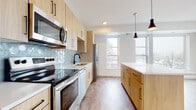 This single-floor space showcases a thoughtfully designed contemporary interior with a cohesive aesthetic throughout. The kitchen serves as a standout feature, offering a galley-style layout on one side with full stainless steel appliances, including a range, built-in microwave, dishwasher, and refrigerator. Light wood flat-front cabinetry runs floor to ceiling, complemented by a striking blue hexagonal tile backsplash that adds visual interest. A large central island with a crisp white quartz countertop anchors the space, illuminated by two sleek black pendant lights. The kitchen flows naturally into an open living area, which benefits from multiple large windows that frame outdoor views and fill the space with natural light. A door from the living area provides access to a private exterior balcony. Durable wood-tone flooring runs throughout, tying each area together seamlessly. The bathroom features a clean, minimalist design with a wood-toned floating vanity, white quartz countertop, undermount sink, and a large frameless mirror. Recessed lighting and a neutral color palette contribute to the bright, airy ambiance present throughout the entire unit.