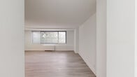 Located at 4921 Seminary Rd, Unit 218, in Alexandria, Virginia, this studio unit offers a thoughtfully organized single-floor layout comprising a living room, kitchen, bathroom, closet, and entrance. The living area is the centerpiece of the unit, characterized by clean white walls, light gray hardwood-style flooring, and a large picture window fitted with horizontal blinds that frames views of surrounding greenery and allows generous natural light to fill the space. A baseboard heating unit runs beneath the window, complementing the bright, airy aesthetic. The open layout provides a flexible canvas for a variety of furniture arrangements. The kitchen is efficiently designed, featuring dark countertops, white cabinetry, and stainless steel appliances including a refrigerator and dishwasher, all set against the same light flooring found throughout the unit. The bathroom includes a full bathtub and shower combination with clean white surrounds. A dedicated closet and a defined entrance area complete the unit's functional layout. The overall ambiance is clean, minimal, and modern, with a consistent neutral palette of white walls and light flooring creating a cohesive and inviting atmosphere throughout.