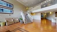 The lobby of Montgomery White Oak Apartments makes a striking first impression with its soaring double-height ceilings and an open, airy atmosphere. Warm hardwood flooring with a decorative inlay pattern stretches across the expansive ground level, anchoring the space with a sense of elegance and comfort. A prominent central staircase with concrete treads and crisp white metal railings serves as the architectural centerpiece, connecting the ground floor to the second-level mezzanine above. The mezzanine overlooks the main lobby area through clean white balustrades, reinforcing the sense of vertical openness throughout.

Textured white painted brick walls add an industrial-chic character to the interior, contrasting beautifully with the warm tones of the flooring. Framed artwork adorns the walls, lending a curated, welcoming feel to the space. A concrete bench with a natural stone seat provides a modern seating option alongside more traditional upholstered armchairs positioned near the entrance. Recessed ceiling lighting illuminates the space evenly, complementing the abundant natural light that pours in through the floor-to-ceiling glass entrance doors. Lush potted greenery near the staircase adds a refreshing natural element. The overall ambiance is polished yet approachable, reflecting a well-designed residential community common area.