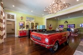 This single-story entertainment space in Columbus, Georgia, delivers a richly themed, multi-zone experience designed for socializing and leisure. The main gathering area features warm hardwood floors, yellow shiplap walls, and crown molding, all bathed in recessed lighting complemented by an industrial pendant light. Iconic egg-style swivel chairs arranged on a pink chevron rug face multiple wall-mounted televisions, while vintage signage—including a classic Frontier and Coca-Cola display—reinforces the retro aesthetic throughout.

A standout centerpiece is a custom pool table crafted from a classic red Ford Mustang convertible, positioned beneath a sculptural chandelier near a striking stone fireplace. Large glass doors alongside this area invite natural light and views of the outdoors. Adjacent to the game room, a fully equipped bar area showcases bold red cabinetry, stainless steel countertops, draft taps, a retro turquoise refrigerator, and a generous high-top table with industrial bar stools.

The screening room offers a distinctly immersive atmosphere, featuring artificial turf flooring, a large projection screen, built-in bench seating with shuttered window accents, and bold chalkboard wall art. Contemporary lounge chairs complete the cinematic setting, making this space equally suited for private movie nights or lively group gatherings.