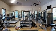 Located at 15 Brasseler Blvd in Savannah, Georgia, this fitness center offers a well-equipped, single-floor workout facility designed to accommodate a comprehensive range of exercise needs. The space features soaring vaulted ceilings that lend an open, airy feel, complemented by multiple ceiling fans that help maintain comfortable airflow throughout. Natural light streams in through several wood-framed windows along the walls, creating a bright and inviting atmosphere with views of surrounding greenery. The interior walls are lined with large mirrors, enhancing the sense of space and allowing users to monitor their form during workouts. The strength training area is well-stocked with a full dumbbell rack, a barbell squat rack with weight plates, and a multi-function cable and weight machine. On the cardio side, treadmills and an elliptical trainer are positioned along the windowed wall, making for an energizing workout experience. The flooring is a durable, easy-to-maintain surface suited for high-traffic fitness use. The overall ambiance is clean, functional, and welcoming, making this facility a practical and comfortable destination for fitness enthusiasts of all levels.