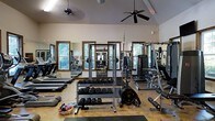 Located at 15 Brasseler Blvd in Savannah, Georgia, this fitness center offers a well-equipped, single-floor workout facility designed to accommodate a comprehensive range of exercise needs. The space features soaring vaulted ceilings that lend an open, airy feel, complemented by multiple ceiling fans that help maintain comfortable airflow throughout. Natural light streams in through several wood-framed windows along the walls, creating a bright and inviting atmosphere with views of surrounding greenery. The interior walls are lined with large mirrors, enhancing the sense of space and allowing users to monitor their form during workouts. The strength training area is well-stocked with a full dumbbell rack, a barbell squat rack with weight plates, and a multi-function cable and weight machine. On the cardio side, treadmills and an elliptical trainer are positioned along the windowed wall, making for an energizing workout experience. The flooring is a durable, easy-to-maintain surface suited for high-traffic fitness use. The overall ambiance is clean, functional, and welcoming, making this facility a practical and comfortable destination for fitness enthusiasts of all levels.