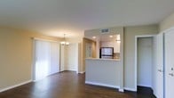 This single-floor space is designed with an open-concept layout that promotes a seamless flow between the living, dining, and kitchen areas. Dark hardwood flooring runs throughout, adding warmth and contrast to the neutral, light-toned walls. The kitchen is positioned behind a raised pass-through counter with a light-colored countertop, offering both a visual divide and a casual breakfast bar. Inside the kitchen, white cabinetry lines the upper walls, complemented by a stainless steel refrigerator and recessed lighting overhead. A stainless steel sink is visible at the counter edge, adding a functional detail. Adjacent to the main living area, a dedicated dining space features an elegant chandelier, providing a focal point for the room. Along one wall, built-in open shelving offers display and storage opportunities. Vertical blind window treatments allow for adjustable natural light. HVAC vents are integrated into the ceiling, ensuring climate control throughout. The overall ambiance is clean, understated, and versatile, suited to a variety of interior styles.