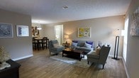 This single-story space offers an open and functional layout that seamlessly connects the living and dining areas. The living room is anchored by a plush gray sectional sofa accented with purple and neutral throw pillows, complemented by a matching accent chair and a dark wood coffee table resting atop a soft area rug. A bold floral artwork adds a vibrant focal point to the neutral beige walls, while a table lamp provides warm ambient lighting. The wood-style flooring runs throughout, lending warmth and visual continuity to the space. A textured ceiling adds subtle architectural character overhead. Transitioning naturally from the living area, the dining space accommodates a dark wood dining table with matching chairs, illuminated by a pendant light fixture that adds a refined touch. A decorative wall arrangement near the dining area adds personality to the space. A doorway visible from the living area suggests additional rooms beyond, contributing to the overall sense of a well-organized and practical floor plan. The overall ambiance is comfortable, modern, and inviting.