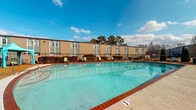This outdoor pool facility offers a well-maintained aquatic space framed by a classic brick-edged pool deck and enclosed by black iron perimeter fencing, providing both safety and a defined boundary for the area. The pool itself is notably spacious, filled with clear, blue-tinted water and clearly marked with depth indicators along its edges. Surrounding the pool, a paved deck provides ample space for lounge chairs, offering residents or guests comfortable spots to relax under open skies with views of mature trees in the background. A multi-unit residential building forms the backdrop, with evenly spaced windows and individual entry doors facing the pool area, reinforcing the communal nature of this amenity. Adjacent to the pool is a dedicated utility and storage shed constructed with wood framing and OSB sheathing. Inside, the shed houses the pool's mechanical systems, including a sand filter tank, pump assembly, and a network of PVC and flexible blue hoses for water circulation and maintenance. Pool rules signage is posted prominently within the shed. Additional storage within the shed includes cleaning tools such as poles and brushes, chemical supplies, buckets, and shelving stocked with maintenance materials and epoxy products, ensuring all necessary upkeep equipment is conveniently on hand.