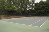 This outdoor tennis court is a dedicated recreational amenity set within a residential community. The court features a green hard surface with clearly marked white boundary lines, fully enclosed by black chain-link fencing that provides both safety and definition to the space. A gated entry point offers convenient access to players. The court surface shows visible cracking, reflecting its established character and history of use.

Surrounding the court is a natural backdrop of tall, mature trees that provide a sense of seclusion and shade. Blooming crepe myrtle trees with vibrant pink flowers add seasonal color and visual appeal to the perimeter. The lush green lawn areas adjacent to the court contribute to an inviting outdoor atmosphere.

Views from within the court look out toward the broader residential community, including multi-story buildings, a well-maintained parking area, and manicured grounds. A light pole is positioned nearby to suggest the potential for evening play. The overall setting offers a functional and accessible recreational space integrated seamlessly into the community environment, ideal for tennis enthusiasts of all skill levels.