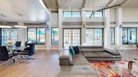 Located on U.S. 98 in Pensacola, Florida, this single-floor commercial space delivers a striking blend of industrial and contemporary design. The main living and lounge area commands attention with dramatically tall ceilings supported by exposed structural beams and trusses, while tiered ring pendant lights and ceiling fans hang overhead. Floor-to-ceiling windows and sets of glass-paned French doors line the walls, flooding the interior with natural light and offering views of the outdoor surroundings. Light hardwood flooring runs throughout, unifying the open layout. Plush gray leather sectional sofas are arranged around a vibrant, multicolored area rug, creating a welcoming and stylish lounge atmosphere. A wall-mounted flat-screen TV and built-in workstation nook with sleek office chairs provide functional workspace alongside a comfortable lounge bench. The reception area near the entrance features a marble-topped desk, modern accent chairs on a teal rug, and a sense of openness accentuated by vaulted ceilings. The galley-style kitchen is well-appointed with white shaker cabinetry, dark countertops, stainless steel fixtures, and ample workspace on both sides. A café bar area, branded as the Cafe Bar of Nottingham, adds a unique hospitality element with a professional coffee station and prep counter, making this space as functional as it is visually impressive.