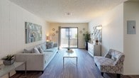 This single-floor residence offers a welcoming and well-appointed living environment designed with comfort and functionality in mind. The living room serves as the focal point of the space, showcasing rich hardwood-style flooring that runs the length of the room, contributing warmth and a sense of continuity. Neutral-toned walls provide a versatile backdrop, complemented by tasteful décor including wall art and decorative accents that add character without overwhelming the space. A large sliding glass door floods the room with natural light and provides direct access to an outdoor balcony or patio area, offering pleasant outdoor views and a seamless connection between indoor and outdoor living. The living area is generously proportioned, comfortably accommodating a full sofa, accent chair, and a sleek glass coffee table. A textured ceiling adds subtle architectural detail overhead, while built-in lighting and wall-mounted fixtures ensure the space is well-lit throughout the day and evening. A white dresser-style console along one wall provides both decorative and functional storage. Overall, the space presents a bright, airy ambiance with a cohesive coastal-inspired aesthetic, making it an inviting and practical home environment.
