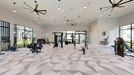 Located at 2300 The Reserve Circle in Apopka, Florida, this single-story fitness center offers a generously proportioned workout facility designed to support a comprehensive range of exercise routines. The main gym area is defined by soaring ceilings adorned with stylish ceiling fans and pendant lighting, creating an open, airy atmosphere that encourages an energizing workout experience. A striking geometric-patterned carpet in neutral tones of white and gray covers the floor throughout, adding a modern, dynamic aesthetic to the space. Floor-to-ceiling windows with sleek black frames line multiple walls, flooding the interior with abundant natural light and offering views of the lush, landscaped grounds featuring palm trees and tropical greenery. The gym is well-equipped with a wide array of machines and equipment, including treadmills, ellipticals, rowing machines, stationary bikes, cable machines, a functional training rig, free weights, and benches. Full-length mirrors are strategically placed to assist with form and enhance the sense of space. The facility also includes a clean, ADA-accessible bathroom featuring neutral tile flooring, a wall-mounted sink, and recessed storage, as well as a utility room. Together, these spaces form a well-appointed, modern fitness destination.