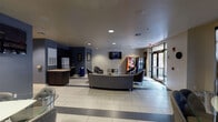 This all-purpose room is a versatile, single-floor communal space designed to accommodate a variety of activities and social gatherings. The layout is thoughtfully organized into distinct functional zones that flow seamlessly into one another across light-toned tile flooring accented by darker border strips.

The lounge area anchors one end of the space, furnished with a large leather sofa, cushioned armchairs, and a glass-topped coffee table with a bold red base. A wall-mounted flat-screen television serves this seating arrangement, making it well-suited for casual viewing or group gatherings. Nearby, a Coca-Cola vending machine and a snack machine offer convenient refreshments.

Toward the opposite end, a dining and activity area features multiple high-top glass tables paired with tufted, adjustable bar-height chairs, providing flexible seating for dining or work. A kitchenette along one wall is equipped with dark cabinetry, granite-style countertops, a stainless steel refrigerator, and a utility sink.

Large floor-to-ceiling windows and glass entry doors line the perimeter, flooding the room with natural light and providing open views of the landscaped outdoor surroundings, including palm trees and walkways. Recessed ceiling lighting and blue accent walls complete the modern, welcoming ambiance.