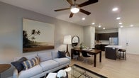 This thoughtfully designed single-floor residence showcases an open-concept layout that effortlessly unifies the living, dining, and kitchen areas into one cohesive and functional space. The living area is anchored by a comfortable light-blue sofa, a glass-topped coffee table, and a large framed coastal artwork that adds a relaxed, inviting ambiance. A ceiling fan with a built-in light fixture and recessed lighting overhead ensure the space remains bright and well-ventilated. The dining area features a rich walnut-toned wooden table paired with a mix of modern black and clear acrylic chairs, creating an eclectic yet sophisticated aesthetic. A round wall mirror and a sleek floor lamp add decorative character to the space. The kitchen, visible from both the living and dining areas, is outfitted with dark espresso cabinetry, light countertops, a subway tile backsplash, and stainless steel appliances, offering both style and practicality. Light wood-tone flooring runs throughout, tying the entire space together with warmth and continuity. The neutral gray wall palette provides a versatile backdrop that complements the modern furnishings and décor, resulting in a space that feels both polished and welcoming.