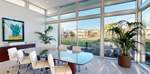 This contemporary office space in Murrieta, CA, showcases a striking blend of modern design and natural elements. The room's most prominent feature is its impressive floor-to-ceiling windows, which flood the space with natural light and offer panoramic views of the surrounding landscape. These windows are complemented by clerestory windows above, enhancing the sense of openness and airiness.

The focal point of the room is a sophisticated glass conference table, accompanied by ergonomic white office chairs. This setup is ideal for meetings and collaborative work sessions. A sleek, mahogany-colored credenza adds warmth to the space and provides essential storage.

Art enthusiasts will appreciate the vibrant, abstract painting featuring bold turquoise hues, which serves as a captivating accent piece. The room's decor is further enhanced by strategically placed indoor plants, including tall palm trees, which bring a touch of nature indoors and improve air quality.

The neutral color palette, dominated by whites and beiges, creates a calm and professional atmosphere. The textured carpet adds comfort underfoot while maintaining the space's modern aesthetic. Recessed lighting in the high ceiling provides additional illumination when needed.

This well-designed office space seamlessly blends functionality with style, creating an environment conducive to productivity and creativity.
