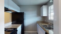 This single-story space presents a cohesive and functional layout finished with consistent design elements throughout. The galley-style kitchen is well-equipped, featuring granite countertops, white cabinetry, and a full suite of black appliances, including a refrigerator, electric range, and dishwasher. A double stainless steel sink sits beneath a window that allows natural light into the space. A standout architectural detail is the pass-through window connecting the kitchen to an adjacent dining or flex area, topped with a granite ledge that adds both utility and visual continuity. This neighboring space is open and versatile, finished with the same light ceramic tile flooring found throughout the unit.

The living areas are generously sized and finished in warm neutral gray tones, with white baseboards providing clean contrast. Textured ceilings run throughout the space, adding dimension overhead. Windows fitted with blinds bring in natural light to the bedrooms, each of which offers a comfortable footprint with tile flooring and ceiling-mounted light fixtures.

The bathroom includes a white vanity with a granite countertop, an undermount sink, and a large wall mirror illuminated by overhead lighting. The overall aesthetic is clean, practical, and move-in ready.