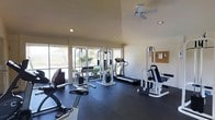 This fitness center offers a comprehensive workout environment designed to accommodate a range of exercise needs. The space features soaring ceilings fitted with recessed lighting and a ceiling fan, ensuring the area remains well-lit and comfortable during use. Large windows line the walls, flooding the room with natural light and providing pleasant views of the surrounding outdoor landscape, including lush greenery and what appears to be a water feature or open grounds beyond.

The flooring consists of durable rubber matting, a practical choice suited for high-impact activities and heavy equipment. The facility is outfitted with a well-rounded selection of exercise machines, including a multi-function cable and strength training system, a treadmill, a recumbent bike, and additional resistance training equipment. A corkboard on the wall provides space for notices or fitness resources. The layout is thoughtfully arranged to allow users to move freely between stations.

The overall ambiance is clean, functional, and inviting, with the abundance of natural light and outdoor views contributing to an energizing atmosphere. The combination of cardio and strength training equipment makes this a versatile space suitable for a full-body workout routine.