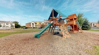 Located at 2332 Asics Dr in Hilliard, Ohio, this single-level outdoor space is home to a well-equipped community playground designed to serve the surrounding residential area. The centerpiece of the space is a substantial wooden play structure finished in warm natural tones, featuring a green slide, a rope climbing wall with colorful hand holds, a tire swing, a covered platform with a teal canopy, and an enclosed playhouse section. The structure is set on a generous bed of wood chip surfacing, providing a safe and cushioned play area underfoot. The open layout allows ample room for movement around the equipment, and the grounds are bordered by well-maintained grass. Surrounding the playground, multi-story brick residential buildings are visible in the background, establishing this as a shared community amenity. The area benefits from open sky exposure, offering an airy and bright outdoor environment. The playground's robust construction and variety of activity stations make it a functional and engaging outdoor destination suited for a range of ages and abilities within the community.