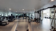 This state-of-the-art fitness center offers a sleek, contemporary workout environment designed to accommodate a wide range of training needs. The facility spans a generously proportioned single-floor layout, providing ample space for members to move freely between equipment stations without feeling crowded.

The interior is finished with a durable, speckled rubberized flooring in a neutral grey tone, ideal for high-traffic athletic use and providing both safety and comfort underfoot. Recessed lighting is evenly distributed across the smooth white ceiling, ensuring consistent, bright illumination throughout the entire space.

The strength training area is outfitted with an impressive array of cable machines, multi-functional cable crossover systems, power racks, and flat utility benches, catering to both beginner and advanced lifters. A full-length mirror wall lines one side of the facility, visually expanding the space while allowing users to monitor their form during workouts.

The open floor plan encourages an organized flow between equipment zones, with clean sightlines across the facility. Large structural columns with a natural concrete finish add an industrial-modern aesthetic contrast to the otherwise crisp, white interior. The overall ambiance is professional, clean, and energizing — a well-considered space built to inspire performance.