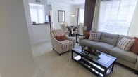 This single-story space showcases an open-concept floor plan that seamlessly connects the living and dining areas, creating a cohesive and functional layout. The living room is anchored by a neutral-toned sofa accented with colorful patterned throw pillows and complemented by a matching accent chair featuring an eye-catching geometric print with warm orange cushions. A sleek glass-top coffee table with dark metal framing serves as a central focal point, adding a contemporary touch to the space.

The flooring throughout is covered in plush, light-toned carpeting that enhances the warm and inviting ambiance. Natural light flows generously into the space through multiple windows dressed with white blinds and sheer curtains, brightening both the living and dining zones.

The dining area, positioned adjacent to the living room, features a dark round table surrounded by upholstered chairs in a neutral tone, topped by an elegant pendant light fixture. A decorative wall-mounted mirror adds depth to the dining space. A dark accent wall provides a bold contrast against the otherwise light color palette. The kitchen pass-through is visible in the background, reinforcing the open and connected nature of the layout.