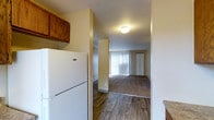 This single-floor residence offers a practical and functional layout designed for comfortable everyday living. The kitchen is equipped with essential appliances, including a full-size refrigerator, and features rich wood-toned cabinetry that adds warmth to the space. Dark hardwood-style flooring runs through the kitchen and transitional areas, providing a clean and durable surface underfoot.

The open sightline from the kitchen leads directly into the living area, which is finished with wall-to-wall carpeting for added comfort. A large vertical blind-covered window in the living space allows natural light to filter in, brightening the interior and creating an inviting atmosphere. The neutral wall tones throughout contribute to a calm and cohesive aesthetic.

Textured ceilings are present throughout the unit, and flush-mount lighting provides consistent illumination. A closet door is visible at the far end of the living area, offering convenient built-in storage. The seamless flow between the kitchen and living zones makes the layout feel open and well-connected, suited for both relaxing and entertaining. Overall, the space presents a straightforward, well-maintained interior with a balance of practical finishes and comfortable details across its single level.
