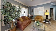 This single-floor professional space is thoughtfully designed to balance functionality with comfort, creating a welcoming environment for both staff and visitors. The reception lounge greets guests with warm, neutral-toned carpeting, slate-blue walls, and crisp white crown molding that lend the space a polished yet approachable character. Plush seating, including a rich brown leather loveseat and patterned gold armchairs, surrounds a decorative glass-top coffee table, while a tall indoor tree adds a touch of natural life. Large windows dressed with horizontal blinds fill the area with natural light and offer views of the surrounding greenery. A skylight further enhances the bright, airy feel of the lounge. An interior pass-through window connects the reception area to the private office, maintaining an open and communicative layout. The private office is generously proportioned, anchored by a substantial wood executive desk and accompanied by two patterned guest chairs. Bold artwork adorns the walls alongside framed credentials, contributing to a distinguished professional ambiance. A multifunction printer and additional workstation support daily operations. Throughout the space, consistent design elements — including the signature blue-gray walls, crown molding, and carpet flooring — create a cohesive and professional aesthetic.