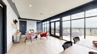 Located at 500 Rolling Mill Hill Rd in Nashville, TN, this rooftop lounge and café space blends Nashville charm with modern design. The interior is defined by deep navy walls, large floor-to-ceiling windows, and light tile flooring that floods the space with natural light. A cozy seating area anchors the main lounge, furnished with plush pink and red accent chairs arranged around a low circular coffee table, set against a striking white pegboard feature wall adorned with an inspirational Dolly Parton quote — a nod to Nashville's iconic culture. A patterned accent wall with a mounted TV and decorative credenza adds visual interest on the opposite side. The café counter area is outfitted with blush-pink cabinetry, a white countertop with a built-in sink, and open shelving displaying a framed Dolly Parton portrait and decorative pampas grass arrangements, reinforcing the space's playful yet sophisticated personality. Expansive glass sliding doors and French doors open directly onto a generous outdoor terrace, where string lights, lounge seating, and dining furniture create an inviting al fresco setting with sweeping views of the Nashville skyline and surrounding landscape. Restroom facilities are conveniently located within the space.