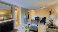 This single-floor residence offers a thoughtfully arranged open-concept layout that combines the living, dining, and entryway areas into one cohesive and functional space. The living area features plush wall-to-wall carpeting and is anchored by a tiled fireplace, adding a warm and inviting focal point to the room. Structural columns subtly define the space without interrupting the open flow between areas. A decorative console table with tall blue vases adds an artistic touch near the entryway corridor. The dining area accommodates a round glass-top table surrounded by sleek black chairs, offering an elegant yet understated setting for meals. The front entry door is clean and simply framed, providing a welcoming threshold into the home. A hallway visible from the main living space leads to additional rooms, including what appears to be a bathroom with distinctive tiled walls. The textured ceiling and warm neutral wall tones throughout create a cozy and consistent ambiance. Overall, the space presents a practical and comfortable single-level floor plan with clearly defined zones for living, dining, and circulation.