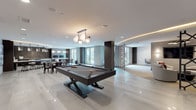 This single-floor amenity space is designed with a sophisticated, contemporary aesthetic that seamlessly connects multiple functional zones. The open-plan layout centers on a game area highlighted by a sleek, dark-framed billiards table set beneath a multi-globe pendant light fixture, with large-format polished tile flooring extending throughout.

An adjacent lounge area features plush white curved seating arranged around a glass coffee table, paired with a wall-mounted flat-screen television and warm chandelier lighting with cove-lit ceiling details. A separate seating alcove showcases a striking three-dimensional textured tile accent wall complemented by starburst flush-mount fixtures overhead and swivel chairs in gray velvet upholstery.

The wet bar and kitchen area is anchored by a curved white countertop island with bar stool seating, dark cabinetry, mosaic tile backsplash walls, and stainless steel appliances. A dining section nearby offers multiple tables and chairs alongside glass doors leading to an outdoor courtyard, allowing natural light to fill the space.

A compact business center provides workstation desks with computers beneath sculptural geometric pendant lights and patterned wallcovering, completing the well-rounded offering of this amenity floor.