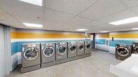 Located at 20-14 Civic Center Dr in East Brunswick, NJ, this commercial laundromat offers a clean, well-organized single-floor facility designed for efficient self-service laundry. The space is immediately recognizable by its vibrant color scheme — bold horizontal stripes of yellow, white, and blue run along the walls, creating a bright and welcoming atmosphere throughout.

The main area is lined with rows of modern, stainless-steel front-load washing machines and dryers, all mounted on raised pedestals for ease of use. The machines are arranged along the perimeter walls, maximizing the open floor space at the center of the room. Large-format light gray tile flooring extends throughout, providing a clean and durable surface that is easy to maintain.

Overhead, a suspended acoustic tile ceiling houses recessed rectangular LED panel lighting, ensuring the space is evenly and brightly lit. Folding tables with attached white seating are centrally positioned, providing customers with a convenient area to sort and fold laundry while they wait. A dedicated utilities room is accessible from the main floor. Windows along the walls allow for some natural light to supplement the interior lighting. The overall layout is practical and functional, with a clear focus on providing customers with a comfortable, efficient laundry experience.