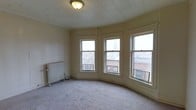 This single-floor space showcases a blend of classic architectural character and urban charm. The most striking feature is a gracefully curved bay window arrangement, comprising multiple large double-hung windows that wrap around the room, flooding the interior with natural light and offering open views of the surrounding cityscape, including rooftops and a distant church steeple. The carpeted floor adds warmth underfoot, while the tiled ceiling lends a period-appropriate aesthetic that speaks to the building's heritage. A vintage cast-iron radiator stands along one wall, providing traditional heating and serving as a nod to the space's historic roots. The neutral, cream-toned walls keep the atmosphere bright and versatile, allowing for a variety of interior design directions. The curved wall following the bay window gives the room a distinctive, non-standard layout that sets it apart from typical rectangular spaces. Overall, the space exudes a sense of timeless urban character, well-suited for those who appreciate architectural detail and the energy of city living.