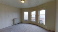 This single-floor space showcases a blend of classic architectural character and urban charm. The most striking feature is a gracefully curved bay window arrangement, comprising multiple large double-hung windows that wrap around the room, flooding the interior with natural light and offering open views of the surrounding cityscape, including rooftops and a distant church steeple. The carpeted floor adds warmth underfoot, while the tiled ceiling lends a period-appropriate aesthetic that speaks to the building's heritage. A vintage cast-iron radiator stands along one wall, providing traditional heating and serving as a nod to the space's historic roots. The neutral, cream-toned walls keep the atmosphere bright and versatile, allowing for a variety of interior design directions. The curved wall following the bay window gives the room a distinctive, non-standard layout that sets it apart from typical rectangular spaces. Overall, the space exudes a sense of timeless urban character, well-suited for those who appreciate architectural detail and the energy of city living.