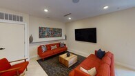 Located at 2142 Commonwealth Ave in Charlotte's vibrant NoDa neighborhood, this single-floor community amenity space at Julien Flats offers a well-appointed gathering area designed for comfort and social connection. The space is anchored by a bright, open lounge featuring bold mid-century modern furnishings, including a pair of striking orange tufted sofas arranged around a rustic wood coffee table atop a patterned area rug. A large wall-mounted flat-screen TV and a vibrant piece of abstract artwork serve as focal points, while recessed lighting and light-toned tile flooring throughout keep the atmosphere clean and welcoming. Floor-to-ceiling glass double doors flood the lounge with natural light and provide views of the outdoor surroundings, reinforcing an open, airy feel. Adjacent to the lounge is a fully equipped kitchenette, thoughtfully designed with granite countertops, white cabinetry, a stainless steel sink, microwave, beverage refrigerator, and a coffee station. Warm wood wall panels add a natural, contemporary contrast to the crisp white walls. A high-top dining table with bar-height chairs and a wall-mounted TV complete the kitchenette area, making it ideal for casual dining or small gatherings. The entrance features a branded entry mat bearing the Julien Flats name, establishing a polished, professional first impression for residents and guests.