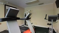This single-floor fitness space is purpose-built for exercise and physical training, offering a focused environment for a full-body workout. The room features a FreeMotion treadmill for cardiovascular training alongside a multi-station cable and strength machine, providing users with both aerobic and resistance training options in one convenient location. A wall-mounted screen or monitor is also present, ideal for viewing workout content or entertainment during sessions. The room is finished with neutral, light-toned walls accented by white crown molding and baseboards, lending a clean and functional aesthetic. Dark carpeted flooring helps define the space as a dedicated workout area while providing cushioning underfoot. Overhead ventilation ensures air circulation throughout the room. The layout is efficient and straightforward, making good use of the available floor space by positioning equipment to allow ease of movement between stations. Overall, the space is practical and well-organized, catering to fitness enthusiasts who value convenience and accessibility in a private or shared gym setting.