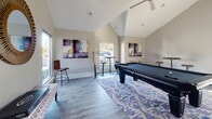 This single-floor recreational space is designed for both entertainment and relaxation, combining thoughtful design elements with a modern aesthetic. The defining architectural feature is the dramatic vaulted ceiling, which rises to a generous peak and creates an airy, open atmosphere throughout the space.

The main area centers around a full-size pool table positioned atop a vibrant blue and white ikat-patterned area rug, serving as the focal point of the room. Tall windows along the walls flood the interior with natural light, offering views of lush outdoor greenery. Wall-mounted flat-screen televisions are positioned for easy viewing, complemented by a plush light gray sofa for comfortable seating.

The décor strikes a balance between bold and refined, featuring a large sunburst gold-framed mirror, abstract artwork in purple, gold, and blue tones displayed across multiple walls, and sculptural gold accent pieces. High-top bistro tables with industrial-style stools provide casual gathering spots throughout the space.

Durable wood-look flooring runs throughout, adding warmth while maintaining a clean, contemporary feel. The neutral wall palette in soft gray-white tones allows the colorful artwork and furnishings to stand out. An entry hallway connects seamlessly to the main lounge and game area.