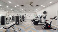 Located at 1623 Parkridge Cir in Crofton, Maryland, this single-floor fitness center is a well-appointed community gym designed to support a comprehensive workout experience. The space is generously sized, offering ample room for both cardio and strength training activities without feeling crowded.

The cardio section features a row of Life Fitness treadmills and elliptical machines, each equipped with individual display consoles and positioned alongside large windows that invite natural light and views of the surrounding greenery. Multiple wall-mounted flat-screen televisions are positioned throughout for entertainment during workouts.

The strength training area is outfitted with a full selection of Life Fitness selectorized machines, a cable crossover station, adjustable benches, and a well-stocked dumbbell rack spanning a wide range of weights. Large floor-to-ceiling mirrors line one wall, enhancing the sense of space and allowing users to monitor their form.

A standout design element is the bold athletic mural painted directly on the white walls, adding an energetic, motivational aesthetic to the space. The flooring features a striking herringbone-patterned carpet in neutral tones with blue accents, lending a modern, polished look. Ceiling fans and recessed lighting ensure the space remains well-lit and comfortable. French glass doors connect to an adjacent area, adding architectural charm to the functional environment.