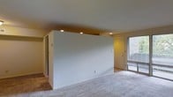 This single-floor residential space offers a clean, functional layout designed for comfortable everyday living. The main living area features soft, neutral-toned carpeting that extends throughout, creating a warm and cohesive feel underfoot. Recessed ceiling lighting provides even, ambient illumination across the space, complementing the bright, white walls that lend a fresh and airy atmosphere.

A central partial-height dividing wall defines distinct zones within the open floor plan, subtly separating areas while maintaining a sense of openness and flow. This architectural detail adds visual interest without sacrificing the spacious feel of the interior.

Near the entryway, a white paneled front door is flanked by a sidelight window, allowing natural light to filter into the space and offering a glimpse of the outdoors. The entry area transitions seamlessly into the main living space, reinforcing the home's open and accessible design.

The overall ambiance is understated and move-in ready, with a neutral palette throughout that provides a versatile backdrop for a variety of interior styles. The thoughtful use of recessed lighting and the balance between defined zones and open space make this a practical and inviting single-level home.
