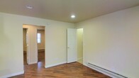 This single-floor space offers a straightforward and functional layout with a clean, neutral aesthetic throughout. The main living area is finished with warm hardwood-style flooring that adds a welcoming character to the space. Smooth, cream-colored walls are paired with white trim and baseboards, creating a bright and airy feel. Recessed lighting in the ceiling provides focused illumination, while baseboard heating units ensure consistent warmth during cooler months.

A six-panel white interior door connects to an adjacent room, which appears to be a utility or bathroom space with tile flooring visible through the doorway. An open passage leads to a secondary room, where a window allows natural light to filter in, enhancing the overall brightness of the unit. Baseboard heating is also present in this adjoining room, confirming consistent climate control across the space.

The layout is open and easy to navigate, with clearly defined rooms connected by wide doorways. The minimalist design and neutral color palette offer a versatile canvas suitable for a variety of uses and furnishing styles. Overall, the space presents a tidy, well-maintained interior with practical features that prioritize comfort and functionality.