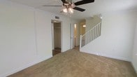 This two-story residence offers a practical and well-organized layout spread across two levels, connected by a classic white-railed staircase that serves as a visual focal point of the main living area. The ground floor features wall-to-wall carpeting in a warm neutral tone, creating a comfortable and cohesive atmosphere throughout the space. Crown molding adds a refined architectural detail along the ceiling line, lending the interiors a polished, traditional character. A ceiling fan with an integrated light fixture is centrally positioned, providing both functional airflow and ambient illumination to the living area. A built-in closet is conveniently located off the main living space, offering practical storage directly accessible from the primary gathering area. Adjacent doorways lead to additional rooms and hallways, hinting at a thoughtful floor plan designed for everyday functionality. The neutral wall tones throughout complement the light-colored trim and baseboards, contributing to a clean and airy aesthetic. The staircase, finished in crisp white with open balusters, rises to the second floor and adds a sense of vertical openness to the interior. Overall, the space presents a straightforward, move-in-ready environment with classic design elements and efficient use of space across both levels.