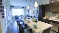 This contemporary single-floor residence showcases a thoughtfully designed open-concept layout that connects the kitchen, dining, and living areas in a seamless, cohesive flow. The kitchen is a standout feature, boasting dark espresso cabinetry with clean-line hardware, a colorful tile backsplash, and a generously sized white waterfall island that doubles as a dining surface. Sleek black dining chairs are arranged along the island, creating a stylish and functional gathering spot. Overhead, elegant pendant lights illuminate the space, complementing the track lighting that runs along the ceiling for versatile ambient coverage. A decorative wall of staggered mirrored panels adds depth and visual interest, reflecting light throughout the open space. The dining area transitions naturally into a comfortable living room bathed in natural light from large windows dressed with flowing white curtains. The living area features a plush seating arrangement anchored by a bold geometric-patterned area rug in blue and white. Soft, neutral wall tones throughout provide a calm and inviting backdrop, while carefully curated décor accents in teal and black tie the entire space together with a polished, modern aesthetic.