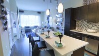 This contemporary single-floor residence showcases a thoughtfully designed open-concept layout that connects the kitchen, dining, and living areas in a seamless, cohesive flow. The kitchen is a standout feature, boasting dark espresso cabinetry with clean-line hardware, a colorful tile backsplash, and a generously sized white waterfall island that doubles as a dining surface. Sleek black dining chairs are arranged along the island, creating a stylish and functional gathering spot. Overhead, elegant pendant lights illuminate the space, complementing the track lighting that runs along the ceiling for versatile ambient coverage. A decorative wall of staggered mirrored panels adds depth and visual interest, reflecting light throughout the open space. The dining area transitions naturally into a comfortable living room bathed in natural light from large windows dressed with flowing white curtains. The living area features a plush seating arrangement anchored by a bold geometric-patterned area rug in blue and white. Soft, neutral wall tones throughout provide a calm and inviting backdrop, while carefully curated décor accents in teal and black tie the entire space together with a polished, modern aesthetic.